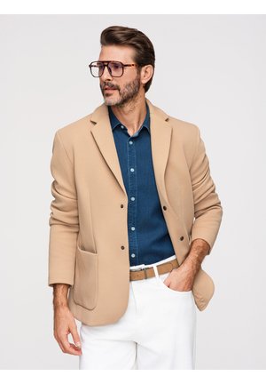 Mężczyzna noszący duże okulary w oprawkach w kolorze skorupy żółwia, beżową marynarkę, niebieską koszulę, białe spodnie i brązowy pasek, pozujący z jedną ręką w kieszeni.