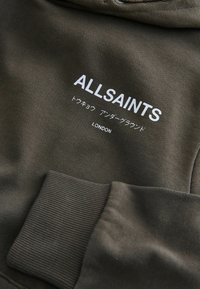 Sweatshirt vert olive avec des poignets côtelés, arborant le texte blanc "ALLSAINTS" et des caractères japonais, avec "LONDON" imprimé en dessous. Texture douce.