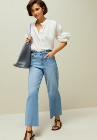 Jeans de pierna ancha azul claro, camisa blanca de botones y sandalias planas marrones. El bolso es gris, de tela, con un diseño holgado.