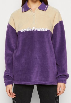 Personne portant un pull polaire violet et beige avec une fermeture éclair sur la moitié et le texte "letsFrans" sur la poitrine.