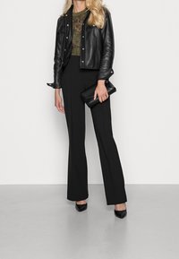 Svart läderjacka över en grön spetsblus, tillsammans med högmidjade svarta byxor och svarta högklackade skor med spetsiga tår, hållande en svart clutchväska.