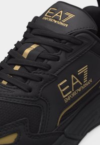 Μαύρο αθλητικό παπούτσι με μαύρα κορδόνια, με χρυσή σήμανση EA7 Emporio Armani στη γλώσσα και στο πλάι, περιλαμβάνοντας μια χρυσή ρίγα στη γλώσσα.