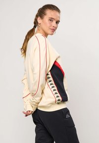 Žena s copom, ktorá má na sebe krémovú mikinu s červeným lemovaním a čiernymi bočnými panelmi s opakujúcim sa čiernym logom, v kombinácii s čiernymi nohavicami.