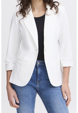 Frau mit einem eng anliegenden weißen Blazer mit gerafften Ärmeln, schwarzem Top und blauen Jeans, die vor einem schlichten Hintergrund steht.