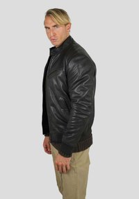 Veste aviateur en cuir noir avec col montant, fermeture éclair à l'avant et deux poches latérales. Ourlet et poignets côtelés ajoutant de la texture. Le mannequin est de profil.