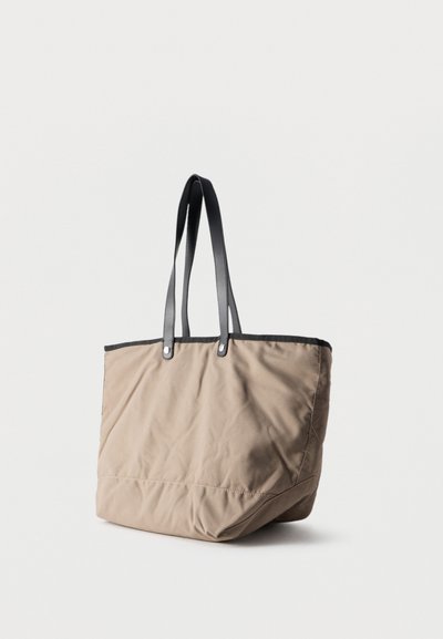 Μπεζ τσάντα υφασμάτινη μαλακή με μαύρες δερμάτινες λαβές και περιθώρια, όρθια σε λευκή επιφάνεια με απλό φόντο.