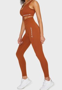 Brązowy top crop i pasujące legginsy z wysokim stanem, wykonane z prążkowanej tkaniny, z logo "SQUATPROOF" w białym kolorze po boku.