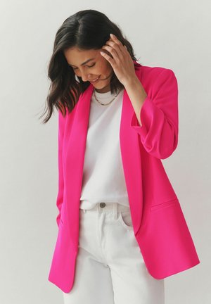 Fuchsiafarbener Blazer mit lockerem Schnitt und hochgekrempelten Ärmeln, kombiniert mit einem weißen T-Shirt und weißen Jeans. Einfaches goldfarbenes Halskettchen als Akzent.