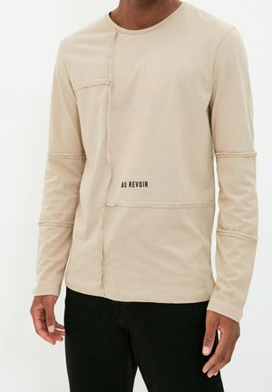 Man draagt een beige overhemd met lange mouwen met zichtbare naden en de tekst "AU REVOIR" boven de taille, gecombineerd met een zwarte broek.