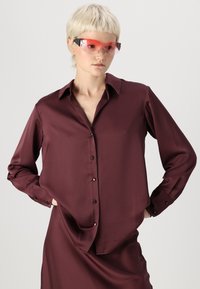 Bordová saténová košeľa s dlhými rukávmi, zapínaním na gombíky a špicatým golierom. Model má na sebe červené slnečné okuliare. Hladká textúra a voľný strih.