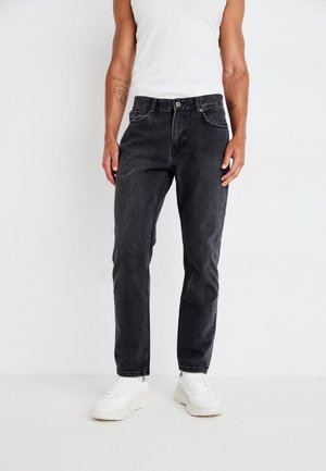 Homme portant un jean droit noir, un débardeur blanc et des baskets blanches épaisses, debout devant un fond blanc uni.