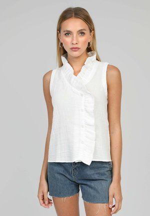 Jeune femme portant un chemisier blanc sans manches à volants et un short en jean bleu, debout devant un fond gris neutre.