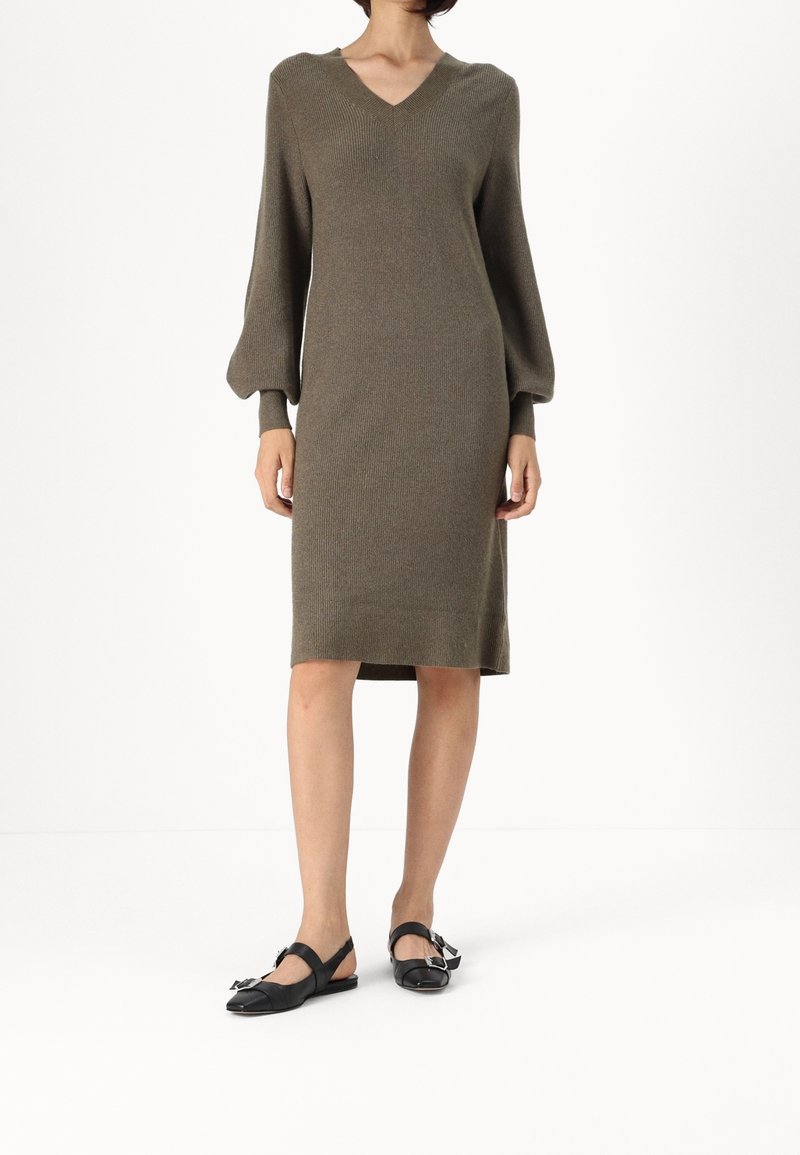 Femme portant une robe en maille vert olive avec un col en V et des manches ballon, associée à des chaussures plates noires à boucle, debout sur fond blanc.