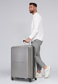 Maleta dura gris con diseño de surcos verticales, mango retractable y cuatro ruedas giratorias, colocada en posición vertical.