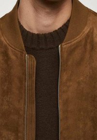 Veste bomber en daim marron avec un col côtelé et une fermeture éclair à l'avant, portée par-dessus un pull en maille marron foncé, mettant en valeur des textures riches.
