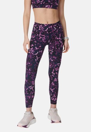 Vrouw draagt donkerpaarse leggings met hoge taille met roze en lavendel bloemmotieven, gecombineerd met lichtroze sneakers en een bijpassende sportbeha.