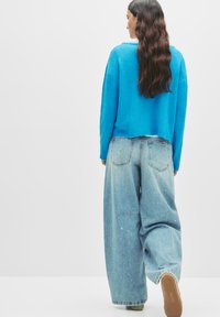 Frau mit langen, gewellten Haaren, die einen leuchtend blauen Pullover und lockere hellblaue Jeans mit Farbspritzern trägt, geht mit einem angehobenen Fuß, mit dem Rücken zum Betrachter.