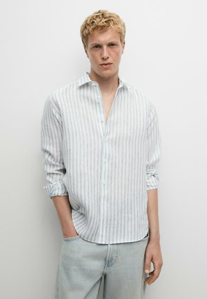 Giovane uomo con capelli biondi ricci che indossa una camicia bianca a maniche lunghe con sottili righe nere e jeans azzurri, in piedi davanti a un muro bianco semplice.