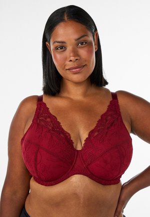 Mulher com pele média e cabelo preto liso veste um sutiã vermelho de renda, sorrindo suavemente com uma mão apoiada na anca contra um fundo simples.