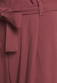 Bordová látka s hladkou texturou, se stáhnutým pasem a jemnými záhyby. Design zdůrazňuje strukturovaný, přesně střižený vzhled.