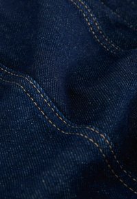 Mörkblå denimtyg med fin väv, som har framträdande gyllene sömmar längs sömmarna, slät struktur och subtila färgvariationer.