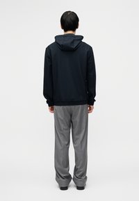 Μπλε καμπαρντίνα με κουκούλα και υφασμάτινη υφή, ελαστικά μανσέτες και τελείωμα· συνδυάζεται με γκρι, φαρδιά παντελόνια. Το μοντέλο φαίνεται από πίσω.