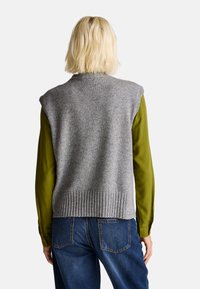 Šedá pletená vesta s ribovaným lemem, nošená přes zelenou blůzu s dlouhým rukávem. Tkanina vypadá texturovaně a zdůrazňuje vrstvený vzhled.