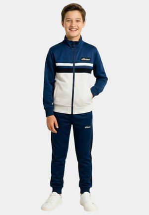 Garçon souriant debout, portant un survêtement zippé bleu et blanc avec des baskets blanches, posant devant un fond clair uni.