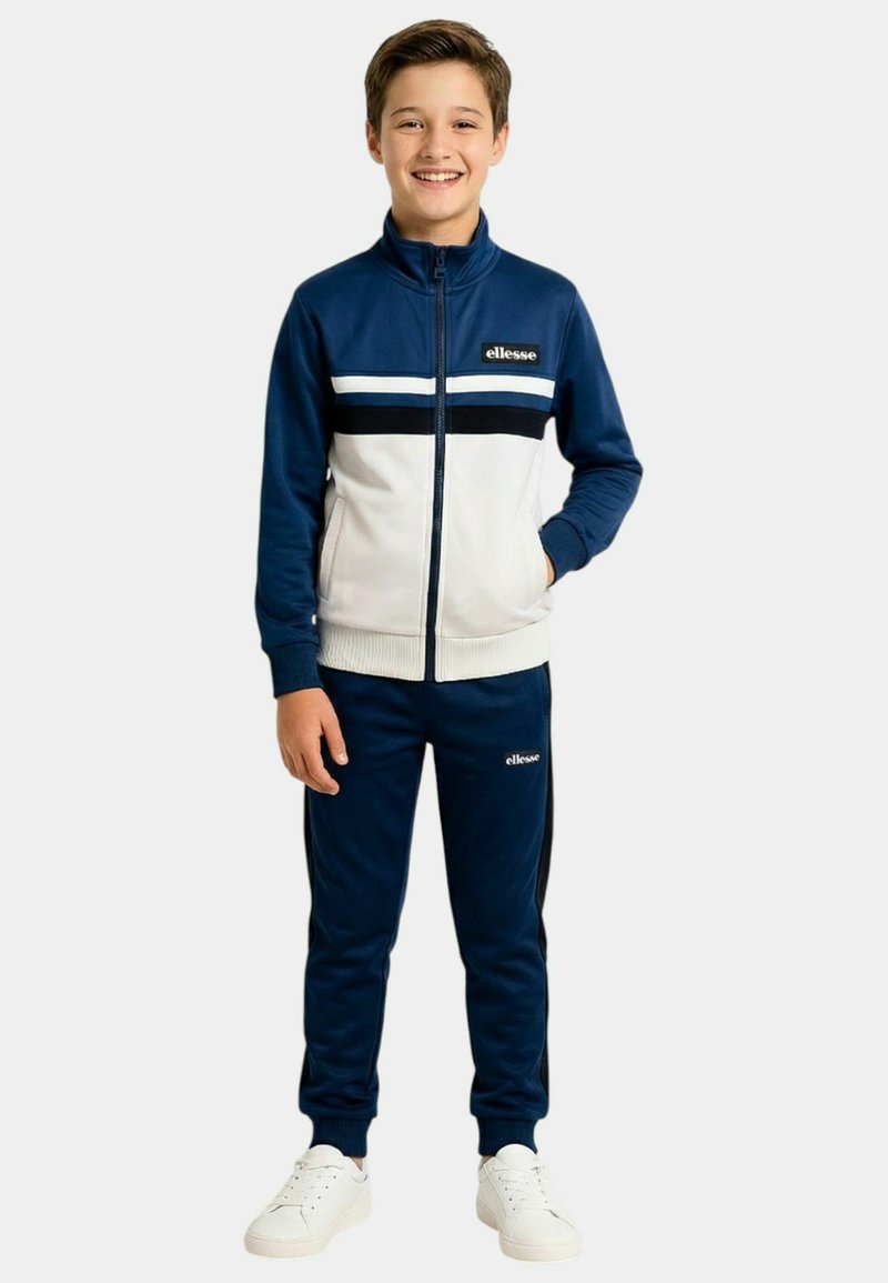 Garçon souriant debout, portant un survêtement zippé bleu et blanc avec des baskets blanches, posant devant un fond clair uni.