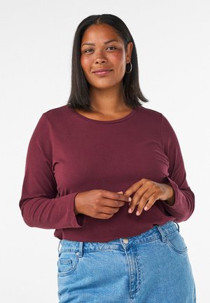 Kvinde med medium mørk hud og skulderlangt sort hår iført en vinrød langærmet trøje og blå jeans, stående mod en hvid baggrund.