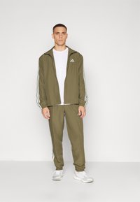 Compleu sport de culoare verde măsliniu cu dungi albe, având o jachetă cu fermoar și mansete elastice. Asortat cu un tricou alb și adidași ușori.