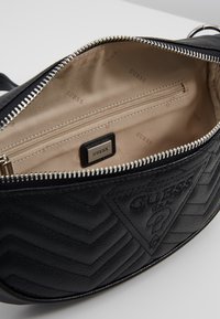 Interno di borsa in pelle nera con rivestimento in tessuto beige, caratterizzato da una tasca con cerniera e un logo "GUESS" discreto. Motivo a chevron sull'esterno.