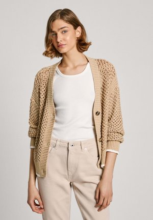Light beige knitted cardigan with open weave and a button closure, worn over a white T-shirt. Casual fit with rolled sleeves.