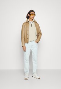Giacca di pelle marrone chiaro, camicia beige con zip sotto, jeans azzurri chiari e sneaker bianche. Occhiali da sole gialli, vestibilità rilassata.