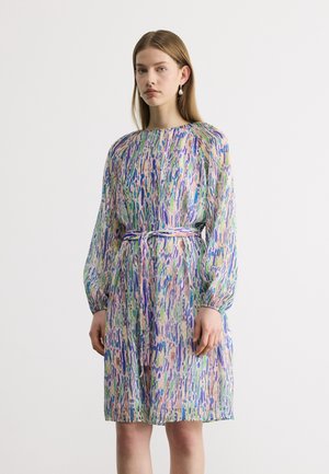 Mlada ženska, oblečena v obleko dolžine do kolen z dolgimi rokavi, s pisanim abstraktnim vzorcem in ujemajočim pasom, zavezanim okoli pasu.