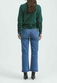 Jeans de tiro alto y pernera acampanada en denim azul con bolsillos traseros, combinados con un suéter verde y elegantes botas de tobillo negras.