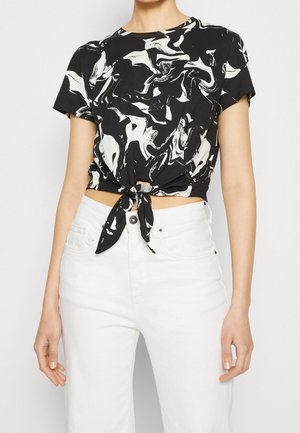 Vrouw in een zwart-wit abstract bedrukt cropped t-shirt met strikvoorpand en korte mouwen, gecombineerd met hoge witte jeans.