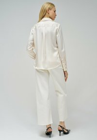 Camisa blanca a rayas de botones con mangas largas, combinada con pantalones anchos blancos y sandalias negras de tacón, mostrada desde la parte de atrás.