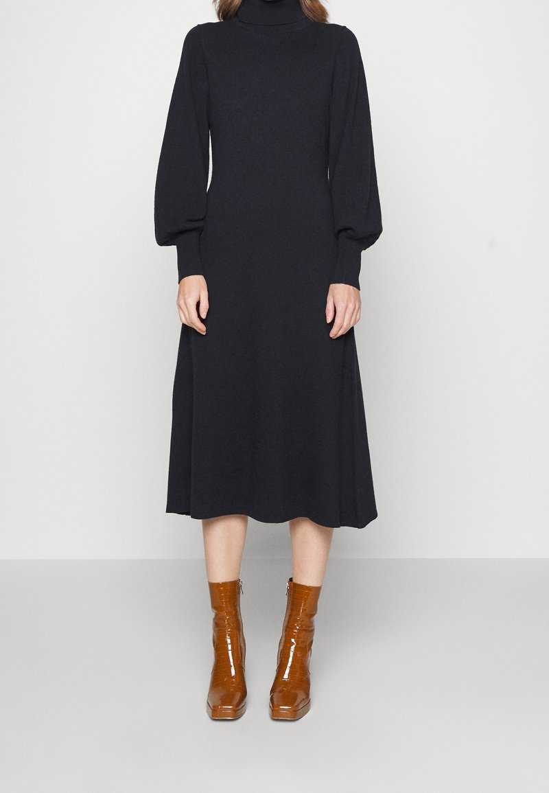 Robe noire à manches longues, longueur genou, avec un col montant imitation col roulé. Texture lisse. Associée à des bottines en cuir verni marron à bout carré.