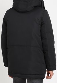 Svart vadderad jacka med huva, långa ärmar och rak skärning. Synlig sömnad och en slät yttre textur.