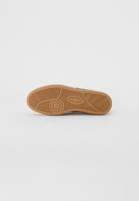 Suola di scarpa in gomma marrone con motivo a zigzag, dettaglio del logo circolare e superficie testurizzata. Progettata per durabilità e trazione.