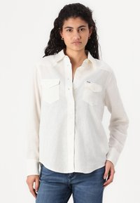 Chemise blanche à manches longues avec boutons en tissu léger, dotée de deux poches poitrine à l'avant et d'un col classique. Portée avec un jean en denim bleu.