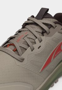 Κοντινή λήψη μπεζ και καφέ παπουτσιού για τρέξιμο σε μονοπάτι με κορδόνια με σχέδιο και κόκκινο λογότυπο "Altra" στο πλάι.