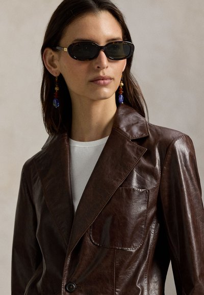 Καφέ δερμάτινο σακάκι με γιακά με χαραγή, ένα κουμπί και τσέπη με μπαλώματα· συνδυασμένο με λευκή ριμπ μπλούζα και μαύρα οβάλ γυαλιά ηλίου.