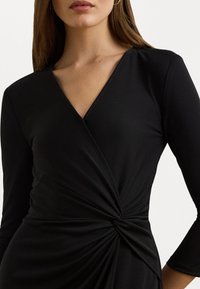 Svart omslagsklänning med långa ärmar, med en V-ringning och tvistdetalj vid midjan. Slät, stretchig tyger med ett elegant utseende.