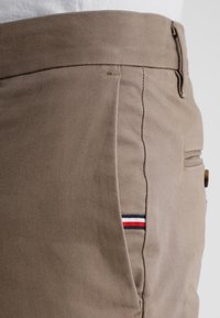 Beige bavlněné kalhoty s úzkým střihem, jemnou texturou a malým červeným, bílým a modrým proužkem na kapse.
