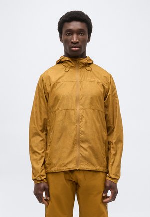 Egy ember, aki mustársárga kapucnis vízálló dzsekit és hozzá illő nadrágot visel, egy egyszínű fehér háttér előtt áll.