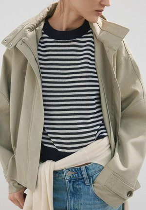 Persoană purtând o geacă bej peste un pulover în dungi alb-negru, cu un pulover bej deschis legat în jurul taliei și blugi albaștri.