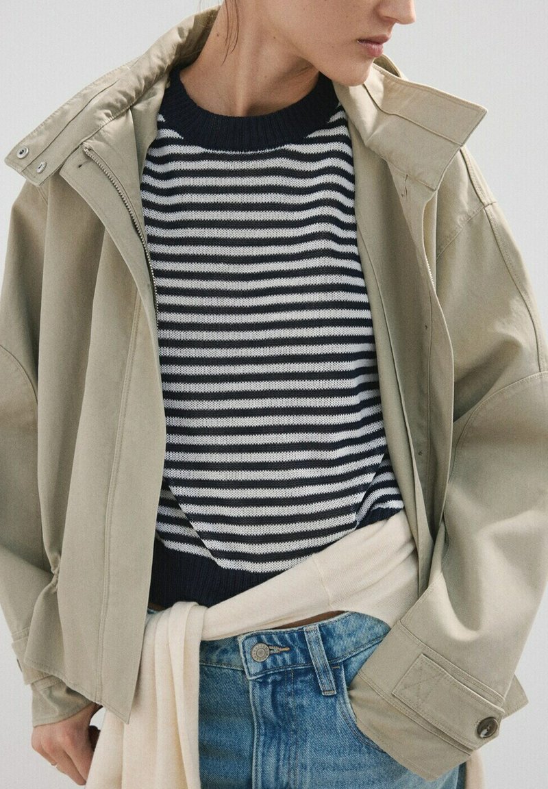 Personne portant une veste beige par-dessus un pull rayé noir et blanc, avec un pull beige clair noué autour de la taille et un jean bleu.