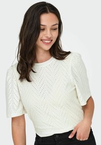 Hvid kortærmet t-shirt med et struktureret, chevron-mønstret udskæringsdesign og puffede ærmer, båret med sorte højhalset bukser.